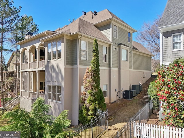 view of home's exterior with stucco siding, fence, roof with shingles, central AC unit, and a chimney