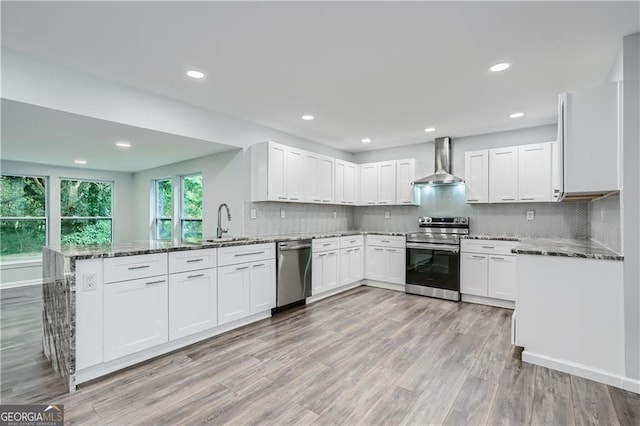 kitchen with a peninsula, dark stone counters, a sink, appliances with stainless steel finishes, and wall chimney exhaust hood