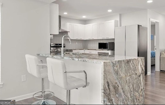 kitchen with tasteful backsplash, a peninsula, black microwave, wall chimney range hood, and light stone countertops