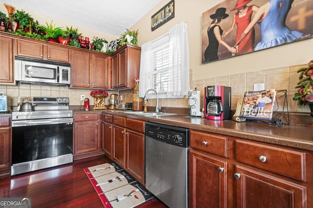 kitchen with dark countertops, a sink, backsplash, stainless steel appliances, and dark wood-style flooring