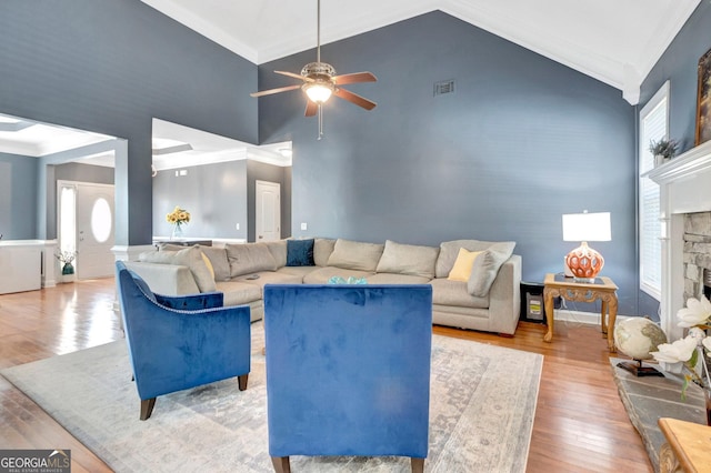 living area featuring high vaulted ceiling, ornamental molding, a ceiling fan, wood finished floors, and a stone fireplace