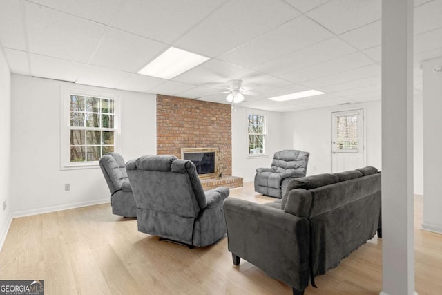 living area with a drop ceiling, light wood finished floors, baseboards, a brick fireplace, and ceiling fan