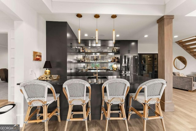 bar with light wood finished floors, hanging light fixtures, stainless steel fridge, and wet bar
