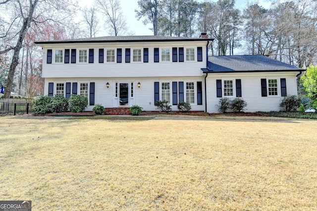 colonial house featuring a shingled roof, a chimney, a front yard, and fence