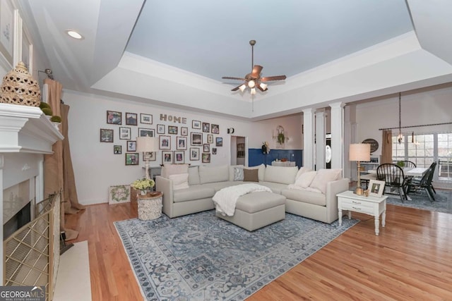 living area with a tiled fireplace, ceiling fan with notable chandelier, light wood finished floors, decorative columns, and a raised ceiling