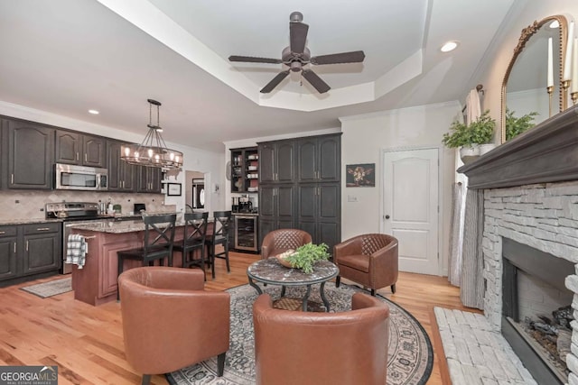 living area with light wood-style flooring, ornamental molding, ceiling fan with notable chandelier, a stone fireplace, and a raised ceiling