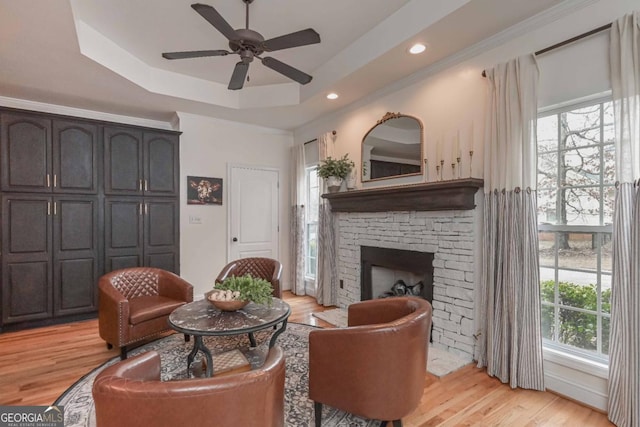 living area with light wood-type flooring, a tray ceiling, a stone fireplace, and a ceiling fan