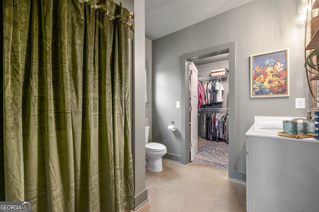 bathroom with a walk in closet, baseboards, finished concrete floors, toilet, and vanity