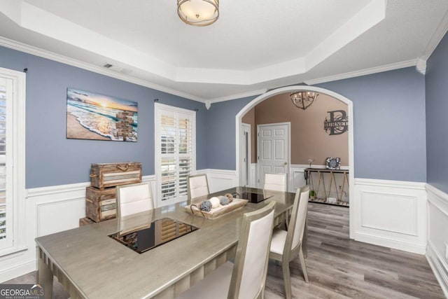 dining space with a wainscoted wall, ornamental molding, a tray ceiling, wood finished floors, and arched walkways