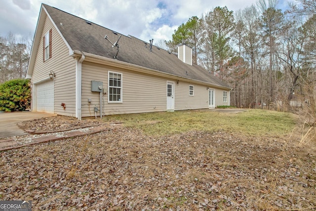 back of house with a shingled roof, concrete driveway, a lawn, a chimney, and an attached garage