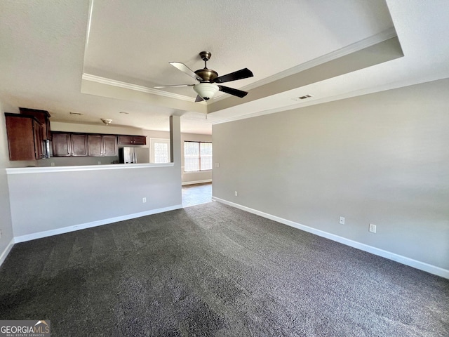 unfurnished living room with crown molding, ceiling fan, baseboards, a tray ceiling, and dark colored carpet