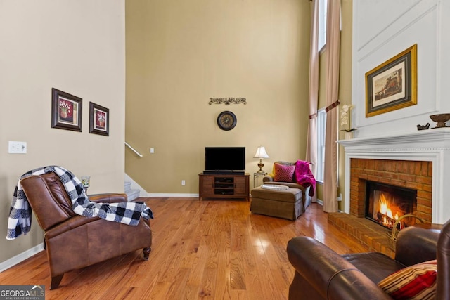 living area featuring a brick fireplace, baseboards, light wood-style floors, and a towering ceiling