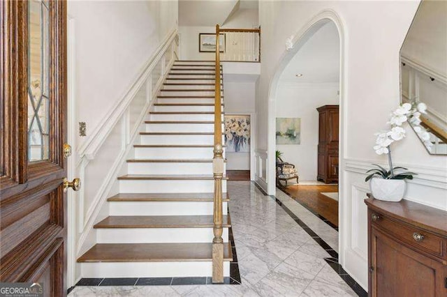 staircase with a high ceiling, arched walkways, crown molding, a decorative wall, and marble finish floor