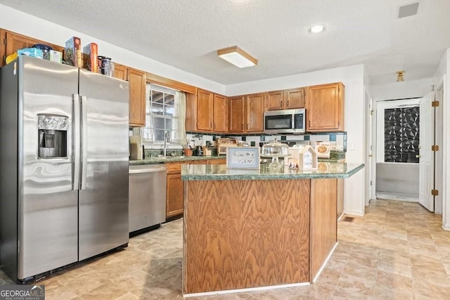 kitchen with brown cabinets, a sink, a textured ceiling, appliances with stainless steel finishes, and a kitchen island
