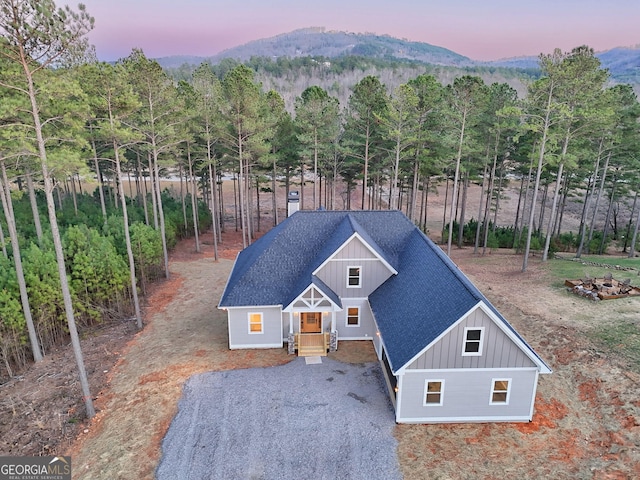 view of front facade featuring a view of trees, a mountain view, gravel driveway, board and batten siding, and a shingled roof