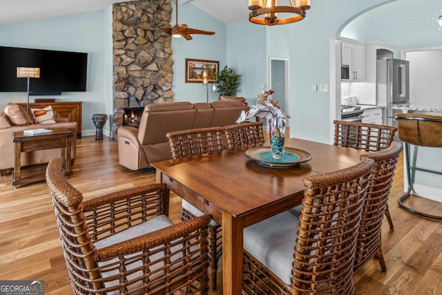 dining space featuring light wood finished floors, high vaulted ceiling, a fireplace, arched walkways, and ceiling fan with notable chandelier