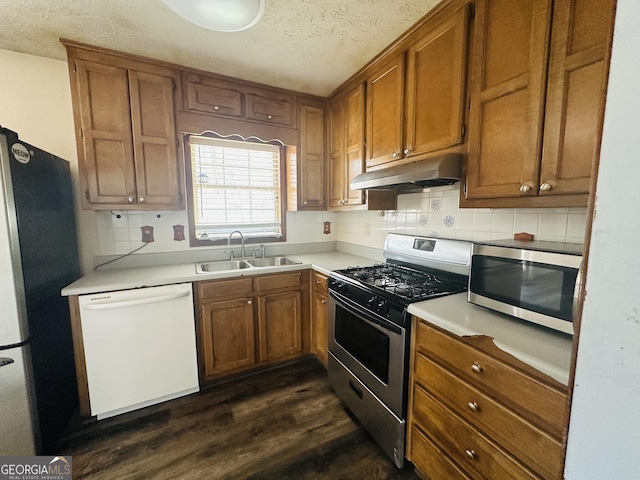 kitchen featuring dark wood-type flooring, under cabinet range hood, appliances with stainless steel finishes, brown cabinetry, and a sink