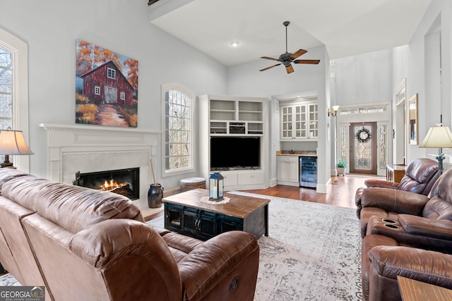 living room with beverage cooler, a glass covered fireplace, wood finished floors, a high ceiling, and ceiling fan