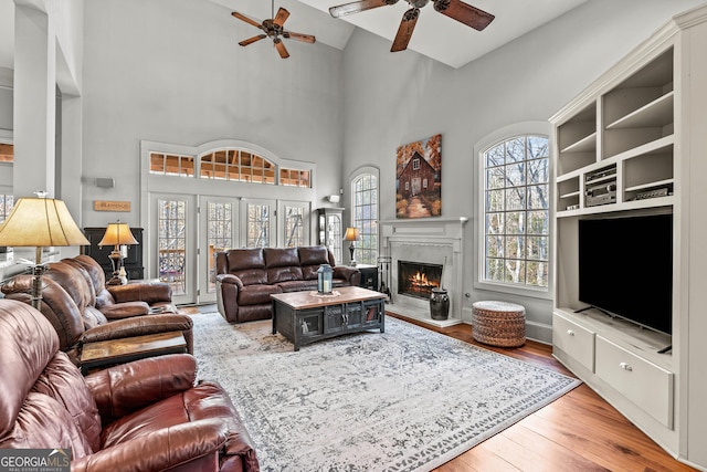 living area featuring a towering ceiling, a warm lit fireplace, wood finished floors, and a ceiling fan