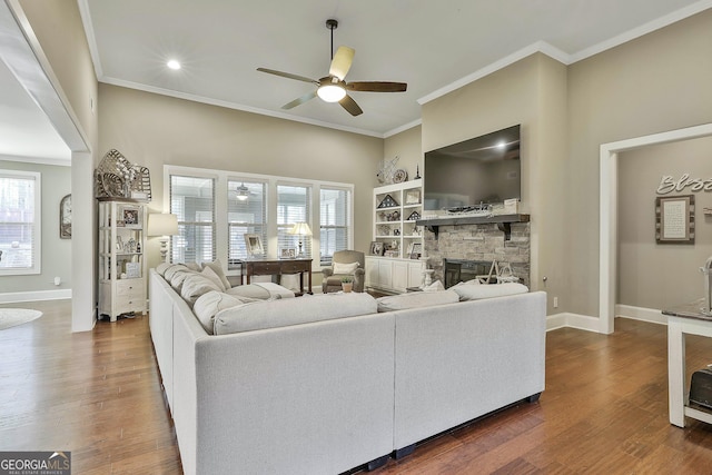 living room featuring dark wood-type flooring, a ceiling fan, a stone fireplace, crown molding, and baseboards