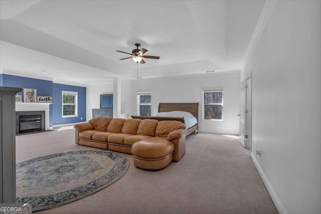 carpeted bedroom with a tray ceiling, a glass covered fireplace, crown molding, baseboards, and ceiling fan