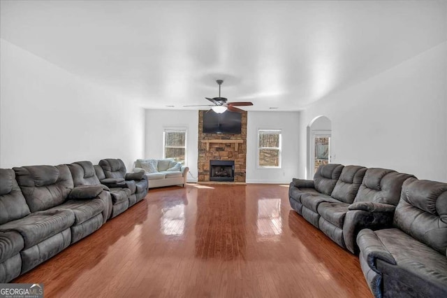 living area with a stone fireplace, wood finished floors, arched walkways, and ceiling fan