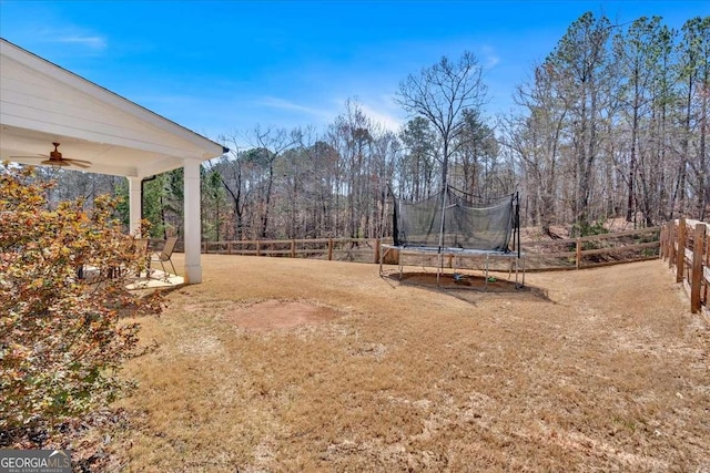 view of yard with a trampoline, a ceiling fan, fence, and a forest view