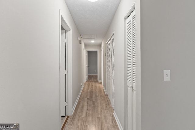 hallway featuring attic access, baseboards, light wood finished floors, and a textured ceiling