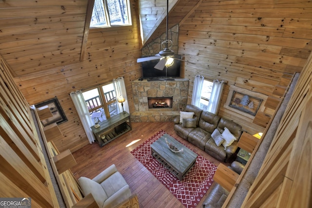 living room with wooden walls, ceiling fan, a stone fireplace, high vaulted ceiling, and wood-type flooring