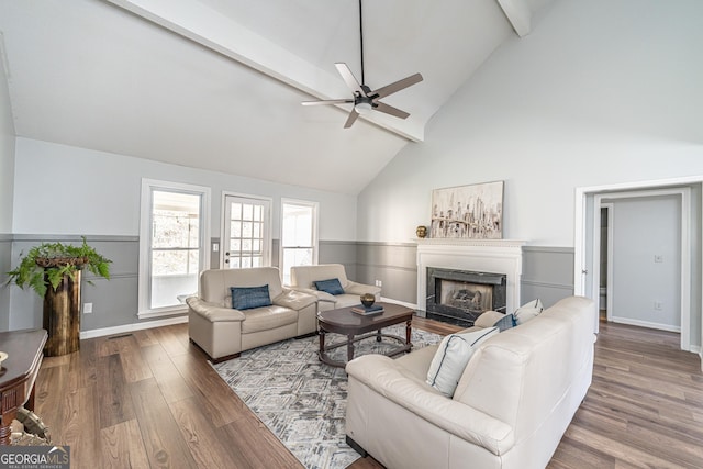 living room with beamed ceiling, a premium fireplace, wainscoting, wood finished floors, and a ceiling fan