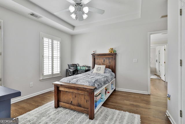 bedroom featuring visible vents, a ceiling fan, a tray ceiling, wood finished floors, and baseboards