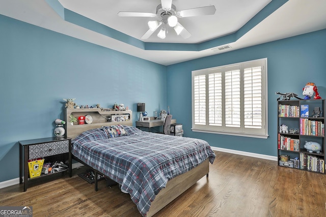 bedroom featuring visible vents, a ceiling fan, a tray ceiling, wood finished floors, and baseboards