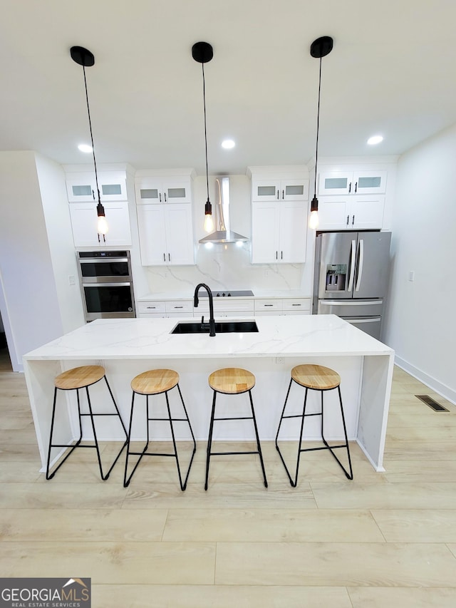 kitchen featuring light wood finished floors, wall chimney range hood, decorative backsplash, stainless steel appliances, and a sink