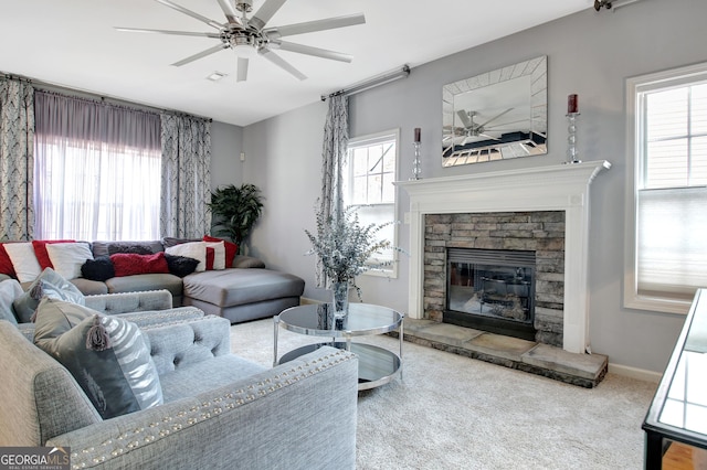 carpeted living area with a wealth of natural light, baseboards, a stone fireplace, and a ceiling fan