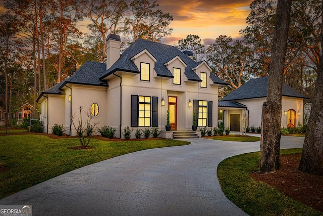 french provincial home featuring a front yard, a shingled roof, stucco siding, curved driveway, and a chimney