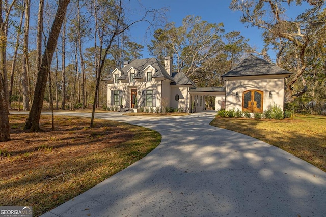 french country inspired facade featuring curved driveway, an attached carport, a front yard, stucco siding, and a chimney