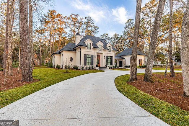 french country style house featuring stucco siding, curved driveway, a chimney, and a front lawn