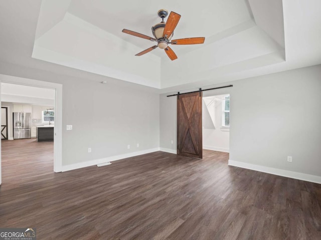 unfurnished room with a tray ceiling, a barn door, baseboards, and dark wood-type flooring