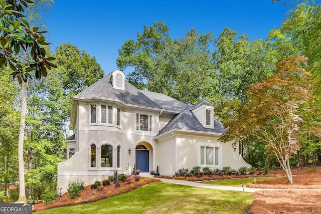 french country style house featuring stucco siding, a front yard, and roof with shingles