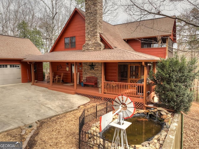 back of house featuring driveway, a porch, a chimney, a garage, and a shingled roof