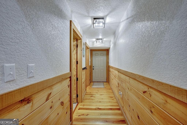 hall with a textured ceiling, wooden walls, wainscoting, light wood finished floors, and a textured wall