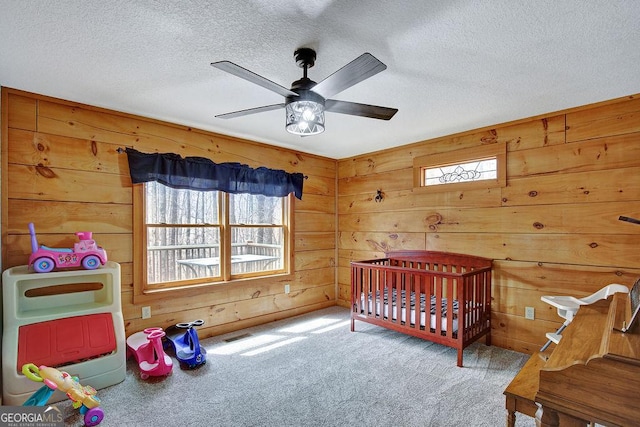 bedroom with a ceiling fan, a textured ceiling, a nursery area, wooden walls, and carpet