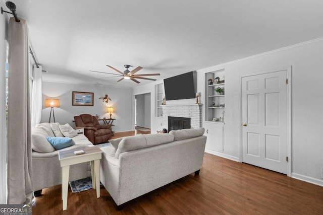 living area featuring built in shelves, baseboards, dark wood-style flooring, ceiling fan, and a brick fireplace