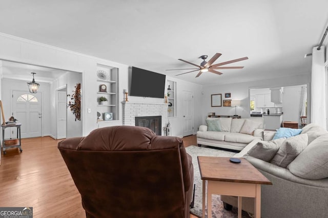 living area with built in features, ceiling fan, a brick fireplace, and light wood-style floors