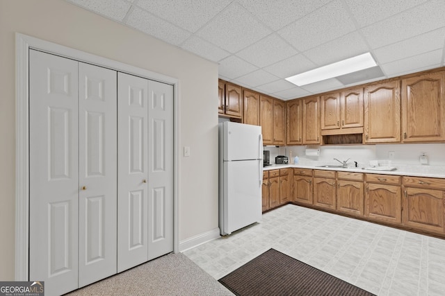 kitchen featuring baseboards, a drop ceiling, light countertops, brown cabinets, and freestanding refrigerator