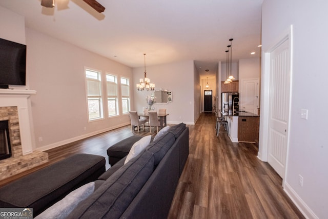 living room featuring baseboards, recessed lighting, a stone fireplace, dark wood-type flooring, and ceiling fan with notable chandelier