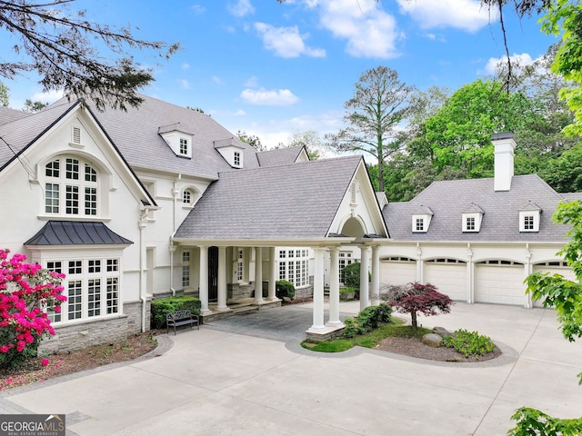 french country inspired facade with a garage, stucco siding, a chimney, covered porch, and driveway