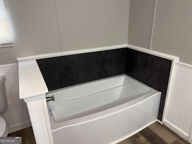bathroom featuring wood finished floors, toilet, a bath, and a wainscoted wall
