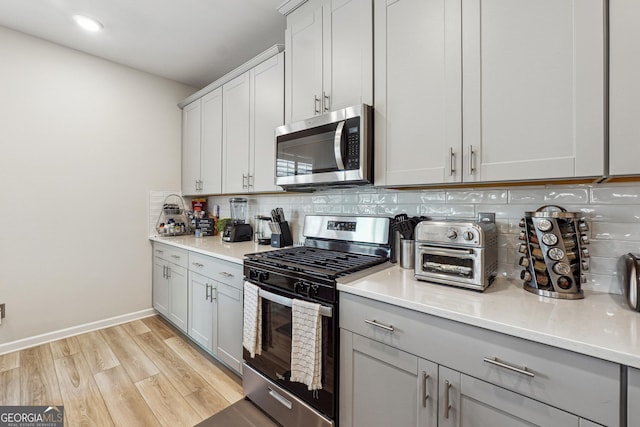 kitchen featuring baseboards, light wood finished floors, gray cabinetry, stainless steel appliances, and backsplash