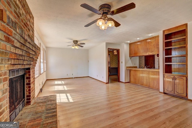 unfurnished living room with light wood-type flooring, a textured ceiling, baseboards, a brick fireplace, and ceiling fan